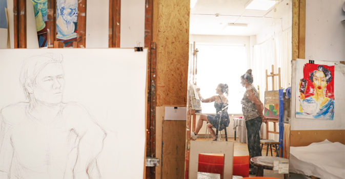 na fotografii pracownia malarska - z lewej fragment szkicu mężczyzny, w środku w lustrze odbiaja się osoba rysująca i istruktorka, z prawej obraz kolorowy przedstawiający portret kobiety