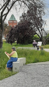 kobieta siedzi na kamiennym murku i rysuje w plenerze, wokół park i ścieżki 