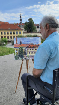siedzący tyłem mężczyzna maluje obraz na sztaludze - budynki miejskie